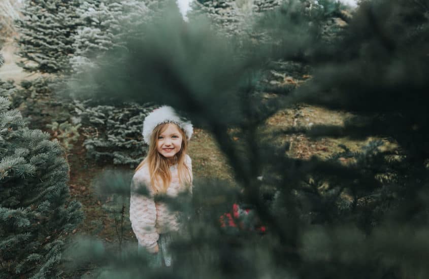 Kleines Mädchen mit Weihnachtsmütze steht zwischen Tannenbäumen