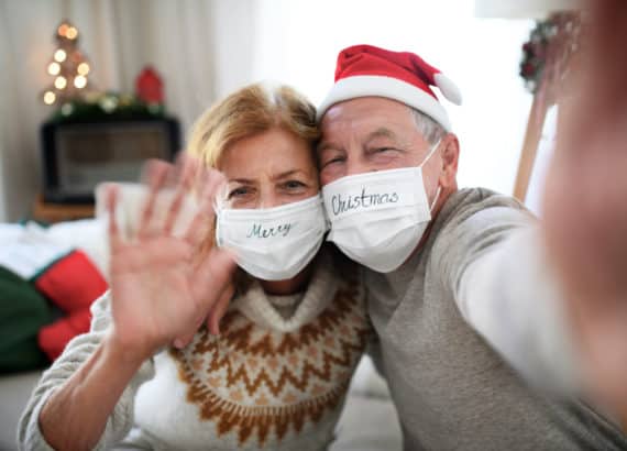 Älteres Pärchen hat Maske und Weihnachtsmützen an und winkt in die Kamera während Weihnachten im Corona Jahr