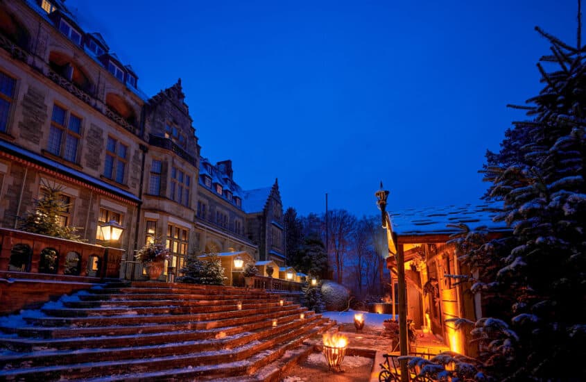 In der Dunkelheit sind die verschneiten Treppen und das Schloss Kronberg festlich erleuchtet. Gegenüber vom Eingang in das Schloss steht eine einladend beleuchtete Weihnachtsmarkthütte.