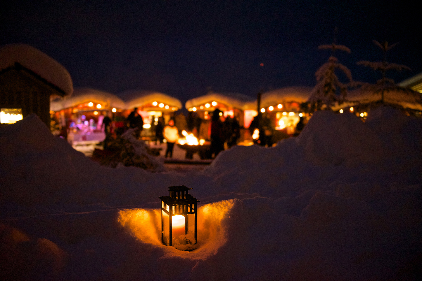 Eine Laterne mit einer brennenden Kerze steht im Schnee, während sich im Hintergrund Menschen das Handwerk und Essen an den Ständen des Weihnachtsmarkts angucken.