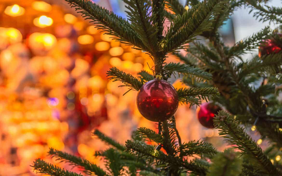 Blick auf die Äste einer grünen Tanne mit roten Christbaumkugeln. Dahinter sind hell erleuchtete Hütten von einem Weihnachtsmarkt.