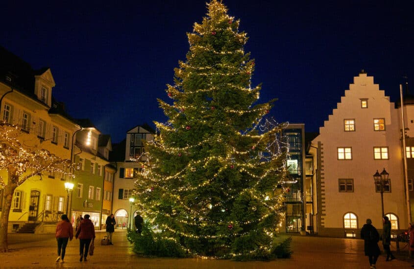 In der Mitte eines Platzes, über den Menschen gehen, steht ein großer Weihnachtsbaum, der festlicht mit Lichtern geschmückt ist.