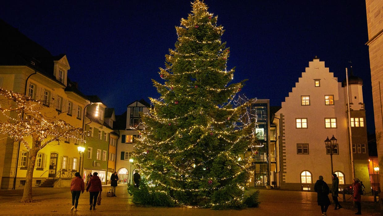 In der Mitte eines Platzes, über den Menschen gehen, steht ein großer Weihnachtsbaum, der festlicht mit Lichtern geschmückt ist.