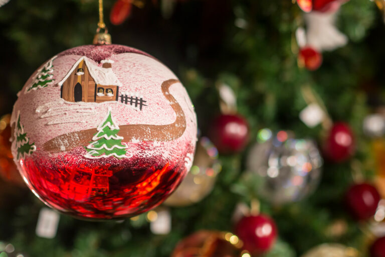 In einem Stand auf dem Weihnachtsmarkt hängt eine bemalte rote Christbaumkugel mit einer schneebedeckten Landschaft, einem Haus und einem Tannenbaum.