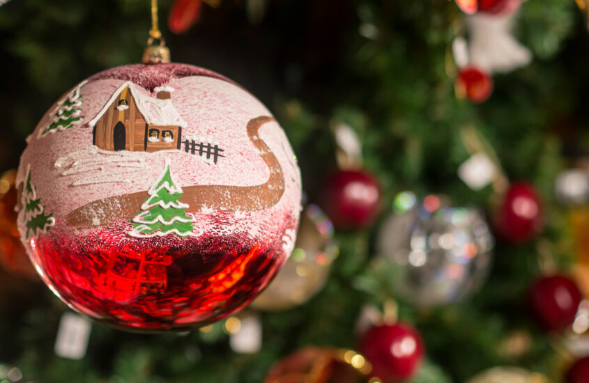 In einem Stand auf dem Weihnachtsmarkt hängt eine bemalte rote Christbaumkugel mit einer schneebedeckten Landschaft, einem Haus und einem Tannenbaum.