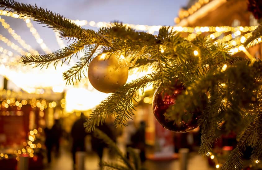 Auf einem hell erleuchteten Weihnachtsmarkt spazieren Menschen zwischen den geschmückten Hütten und grünen Tannenbäumen mit roten und goldenen Kugeln.