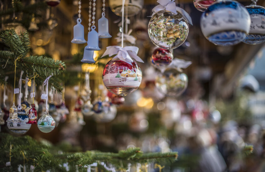 Filigrane Christbaumkugeln aus buntem Glas hängen zwischen Ästen von Tannenbäumen in einem Stand auf dem Weihnachtsmarkt.
