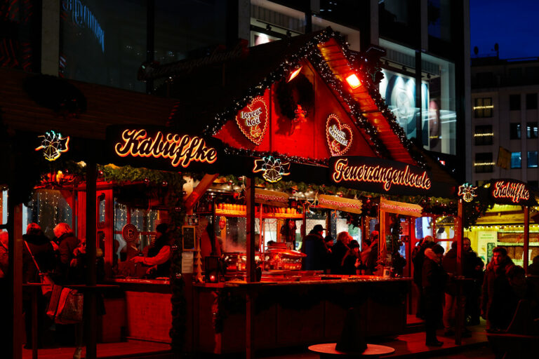 An einer festlich geschmückten Hütte in weihnachtlich rotem Licht auf dem Düsseldorfer Kö-Bogen-Markt lassen sich Besucher ein heißes Getränk schmecken.