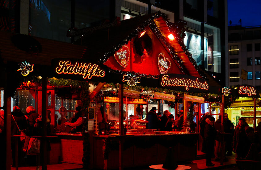 An einer festlich geschmückten Hütte in weihnachtlich rotem Licht auf dem Düsseldorfer Kö-Bogen-Markt lassen sich Besucher ein heißes Getränk schmecken.