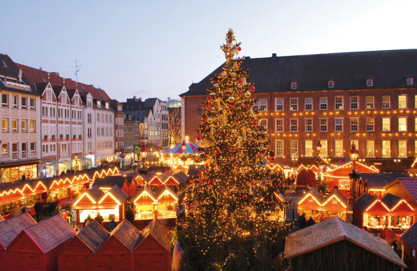 Blick auf den Marktplatz in Düsseldorf und die Hütten des Handwerker-Markts. Alles ist festlich mit Lichtern geschmückt und erleuchtet und in der Mitte steht ein großer Weihnachtsbaum.
