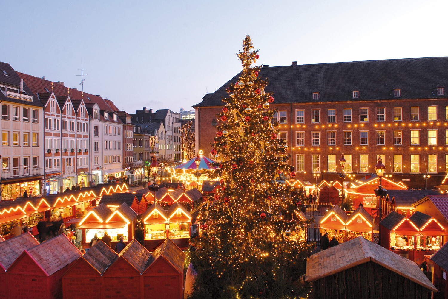 Blick auf den Marktplatz in Düsseldorf und die Hütten des Handwerker-Markts. Alles ist festlich mit Lichtern geschmückt und erleuchtet und in der Mitte steht ein großer Weihnachtsbaum.