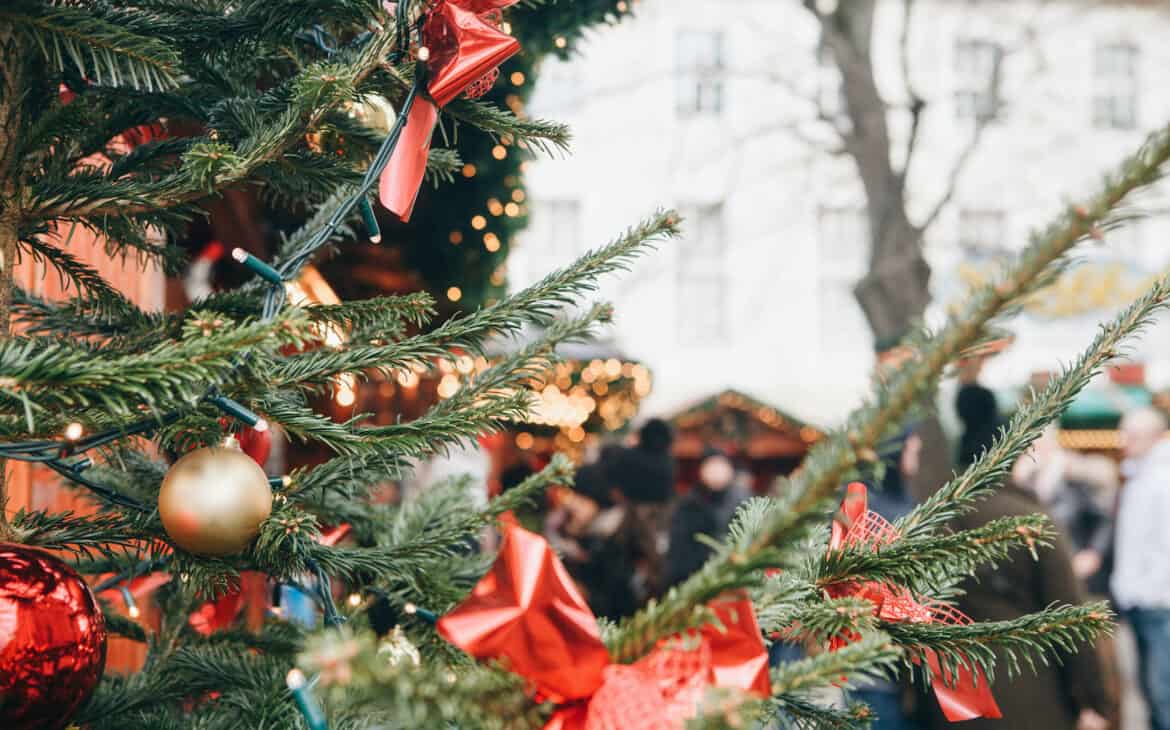 Nahaufnahme von einer mit Kugeln und roten Schleifen geschmückten Tanne auf einem Weihnachtsmarkt in Hamburg. Im Hintergrund sieht man die geschmückten Dächer der Hütten und Menschen, die über den Markt spazieren.