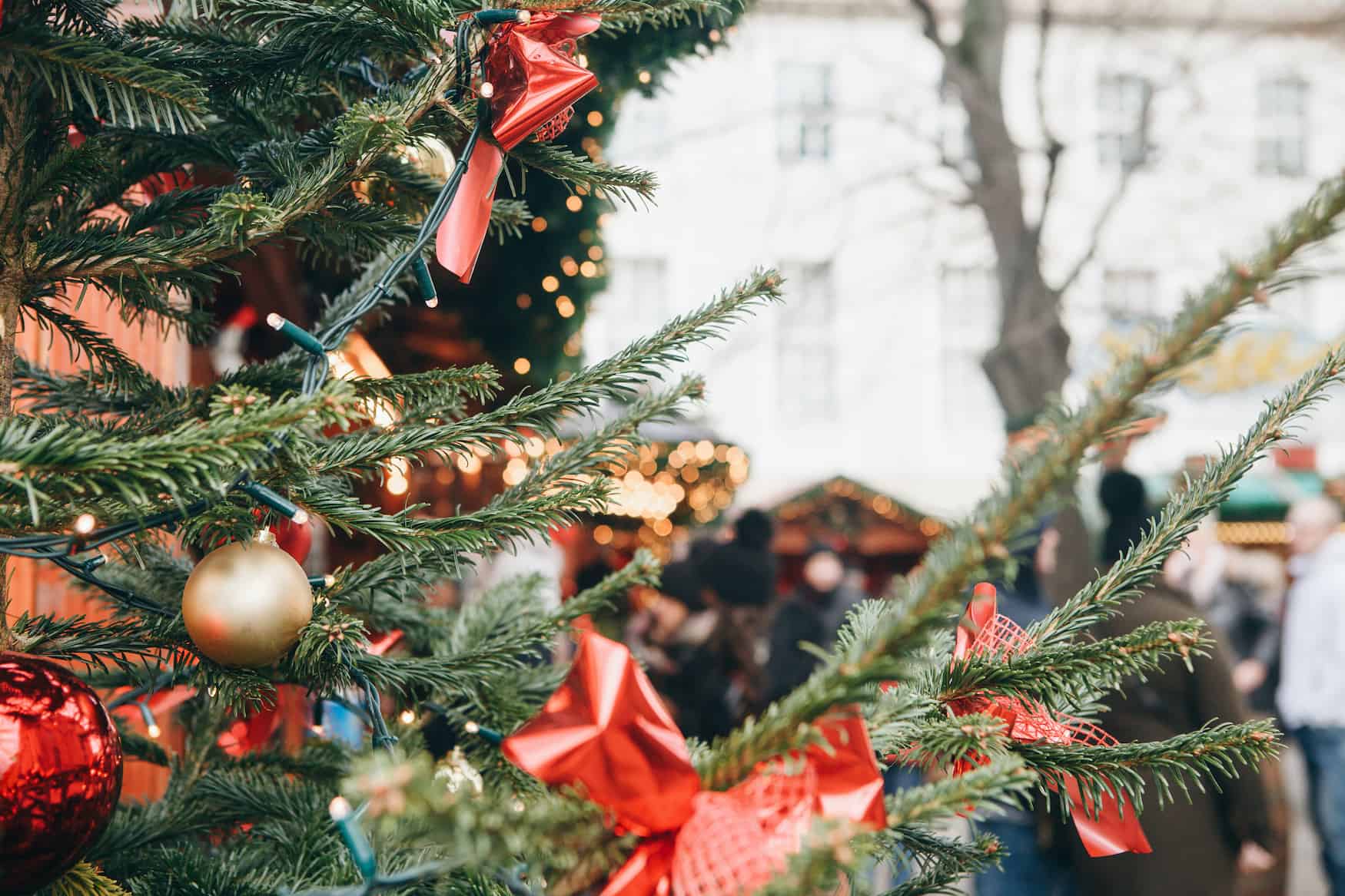 Nahaufnahme von einer mit Kugeln und roten Schleifen geschmückten Tanne auf einem Weihnachtsmarkt in Hamburg. Im Hintergrund sieht man die geschmückten Dächer der Hütten und Menschen, die über den Markt spazieren.