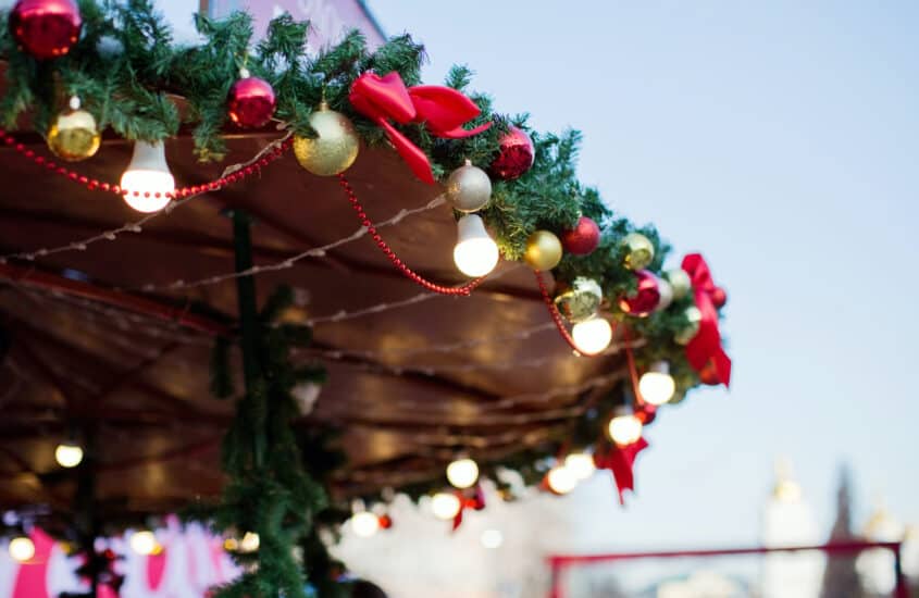 Das Dach einer Hütte auf dem Weihnachtsmarkt ist mit grünen Tannenzweigen, einer hellen Lichterkette und Schmuck aus roten und goldenen Kugeln und roten Schleifen geschmückt.