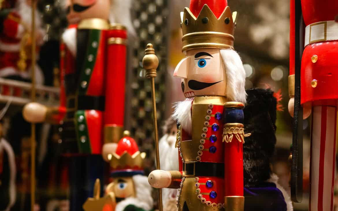 Ein Nussknacker-Soldat in roter Uniform und mit weißem Haar und Bart steht zwischen anderen Nussknackern in einer Hütte auf einem Weihnachtsmarkt.