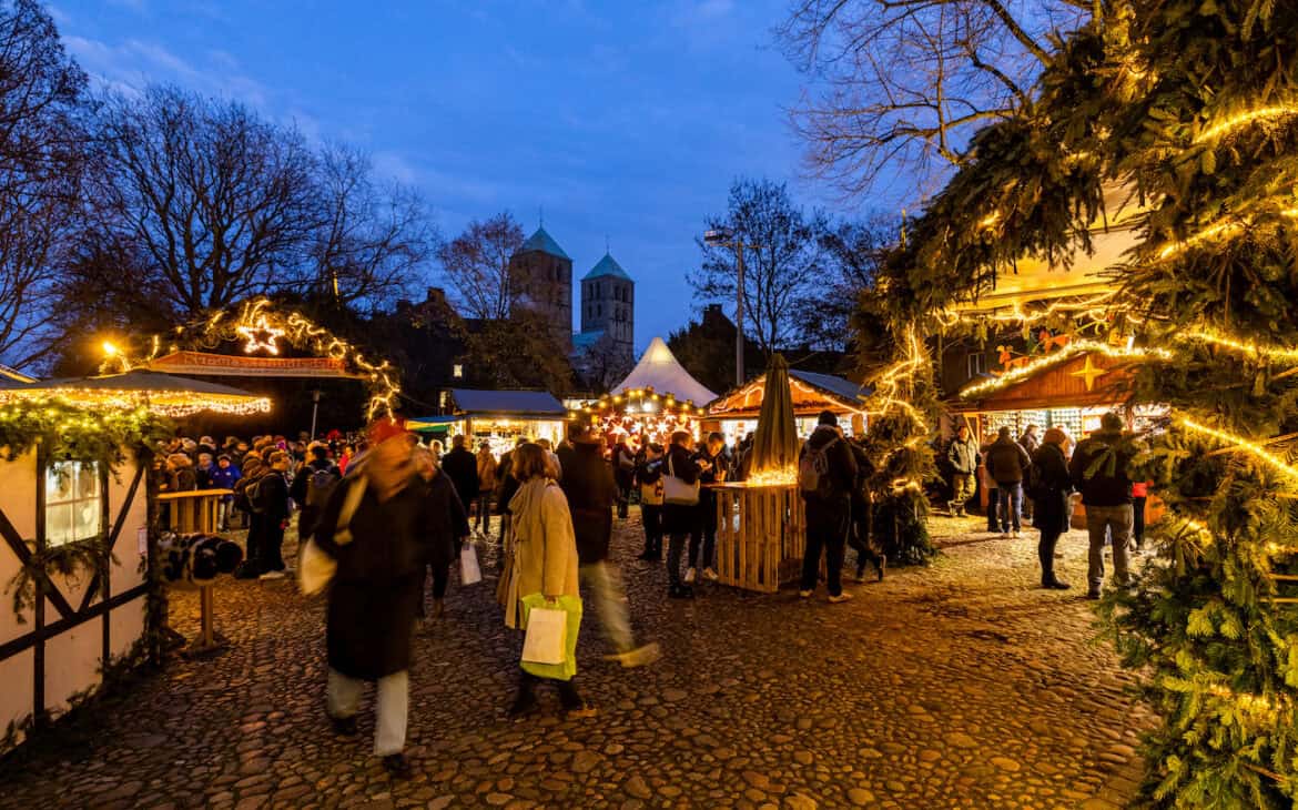 Menschen auf dem Giebelhüüskesmarkt an der Überwasser mit Lichterschmuck und Tannenzweigen, im Hintergrund in der Bildmitte die Türme des Paulusdoms.