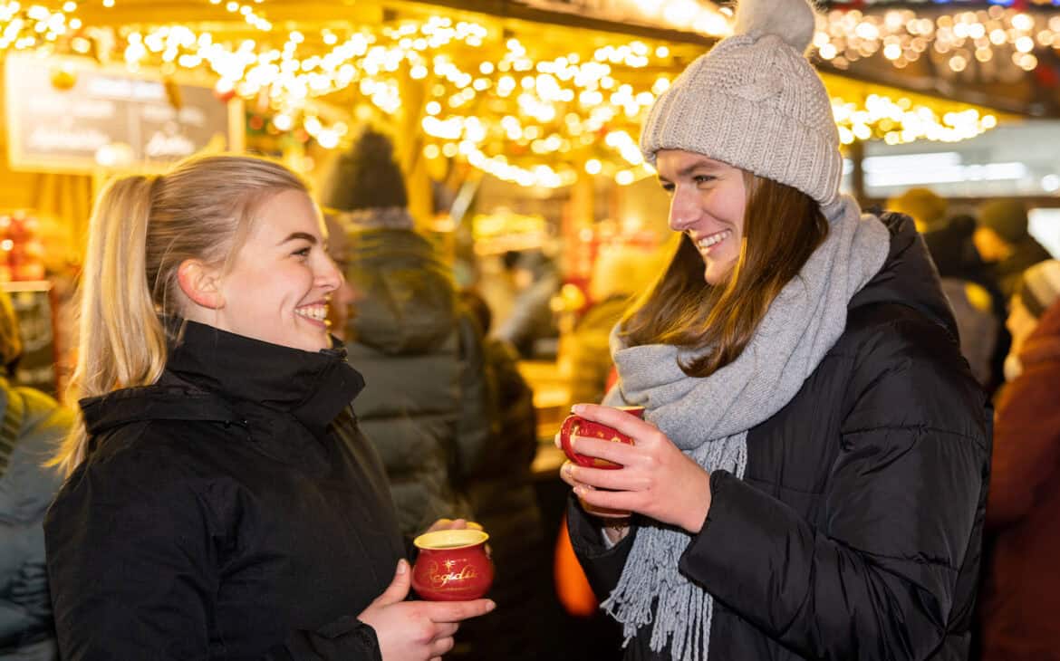 Zwei junge Frauen in Winterkleidung und roten Tassen in den Hand lachen sich an, im Hintergrund festlicher Lichterschmuck des Adventsmarkts.