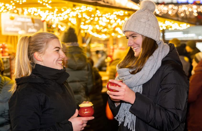 Zwei junge Frauen in Winterkleidung und roten Tassen in den Hand lachen sich an, im Hintergrund festlicher Lichterschmuck des Adventsmarkts.