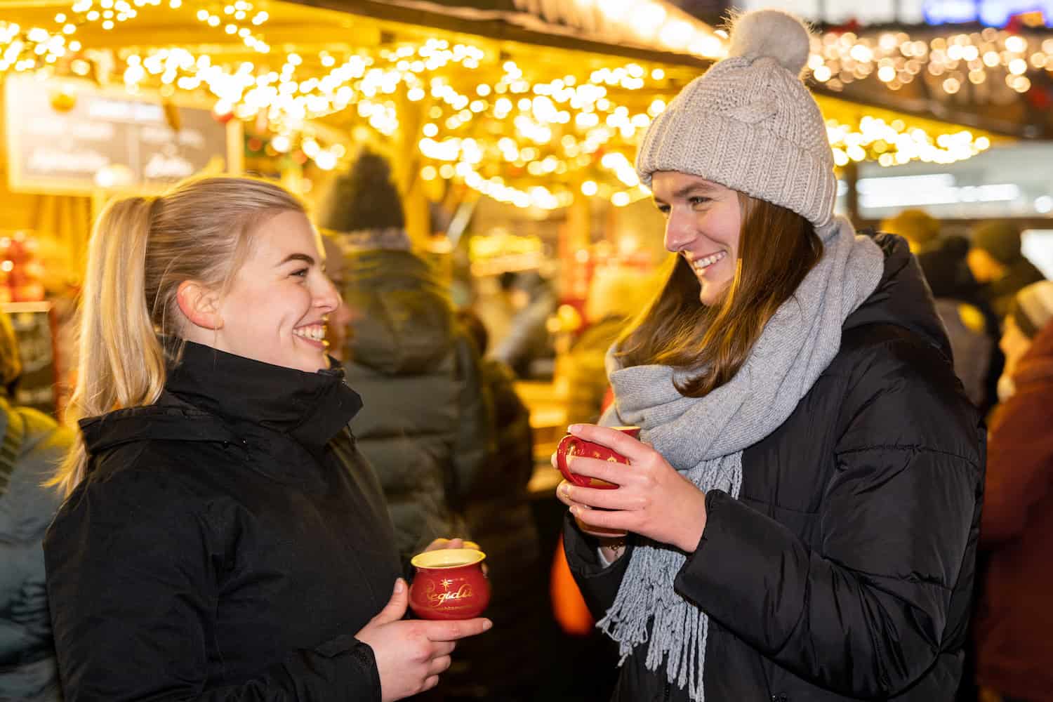 Zwei junge Frauen in Winterkleidung und roten Tassen in den Hand lachen sich an, im Hintergrund festlicher Lichterschmuck des Adventsmarkts.