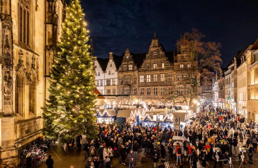 Blick aus leicht erhöhter Position auf den Lambertikirchplatz mit dem Lichtermarkt, festlich beleuchtete Buden, den Weihnachtsbaum an der Lambertikirche und viele Menschen.