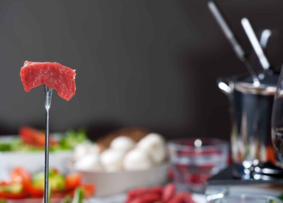 Fleischfondue angerichtet auf Tisch, Fonduegabel mit Fleischstück im Vordergrund