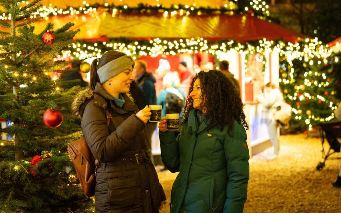 Zwei Frauen in Winterjacken halten auf einem Weihnachtsmarkt Becher in den Händen und lachen sich an, dahinter leuchten Lichterketten an einer Marktbude und geschmückte Tannenbäume.