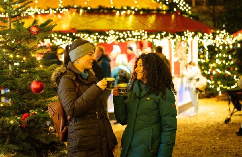 Zwei Frauen in Winterjacken halten auf einem Weihnachtsmarkt Becher in den Händen und lachen sich an, dahinter leuchten Lichterketten an einer Marktbude und geschmückte Tannenbäume.