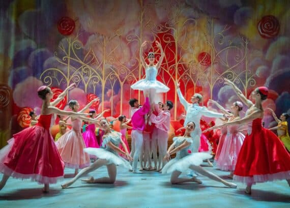 Ensemble des Grand Classic Ballet im Nussknacker: Eine Ballerina im hellblauen Tutu wird vor einem bunten Bühnenbild mit Lollis und goldenen Ranken hochgehoben, Tänzerinnen in roten und rosa Kleidern umringen sie