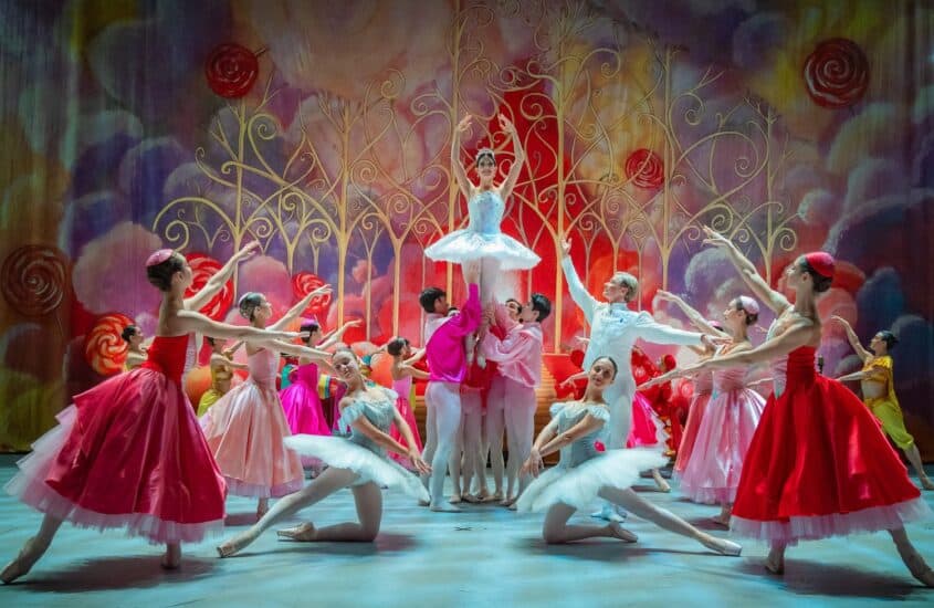 Ensemble des Grand Classic Ballet im Nussknacker: Eine Ballerina im hellblauen Tutu wird vor einem bunten Bühnenbild mit Lollis und goldenen Ranken hochgehoben, Tänzerinnen in roten und rosa Kleidern umringen sie