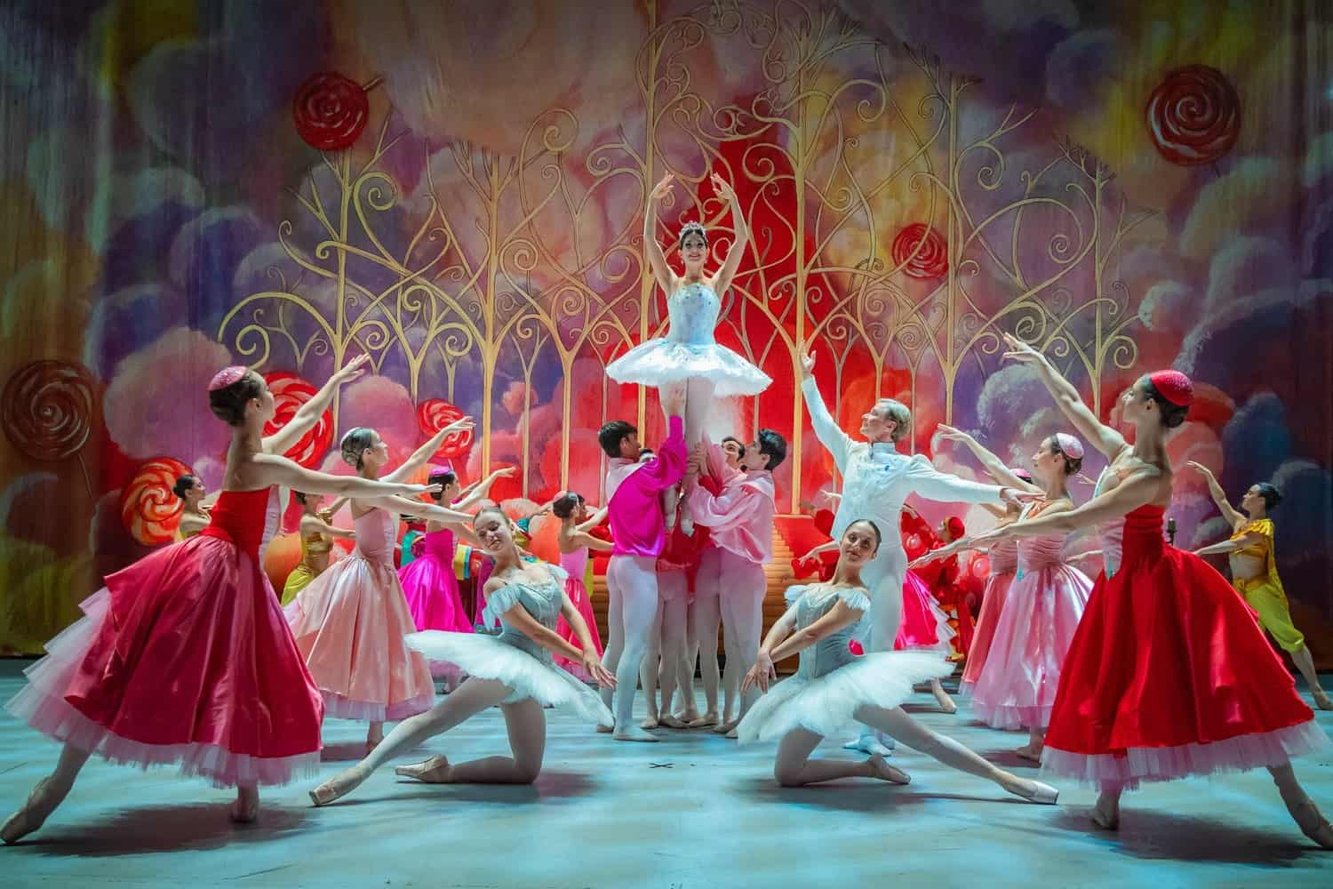 Ensemble des Grand Classic Ballet im Nussknacker: Eine Ballerina im hellblauen Tutu wird vor einem bunten Bühnenbild mit Lollis und goldenen Ranken hochgehoben, Tänzerinnen in roten und rosa Kleidern umringen sie