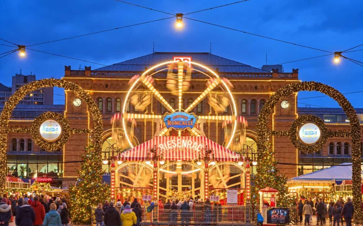 Beleuchtetes Riesenrad im Roncalli Weihnachtsdorf vor dem Hauptbahnhof Hannover zur blauen Stunde, davor Lichterbögen, Weihnachtsbäume und Besucher auf dem Ernst-August-Platz