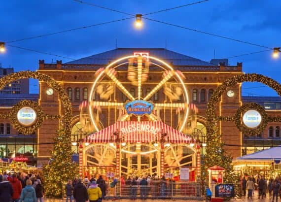 Beleuchtetes Riesenrad im Roncalli Weihnachtsdorf vor dem Hauptbahnhof Hannover zur blauen Stunde, davor Lichterbögen, Weihnachtsbäume und Besucher auf dem Ernst-August-Platz