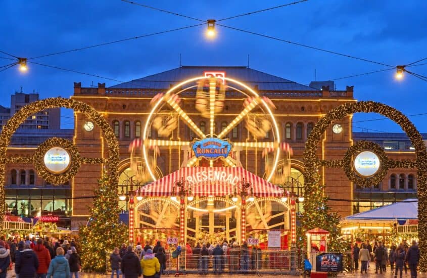 Beleuchtetes Riesenrad im Roncalli Weihnachtsdorf vor dem Hauptbahnhof Hannover zur blauen Stunde, davor Lichterbögen, Weihnachtsbäume und Besucher auf dem Ernst-August-Platz