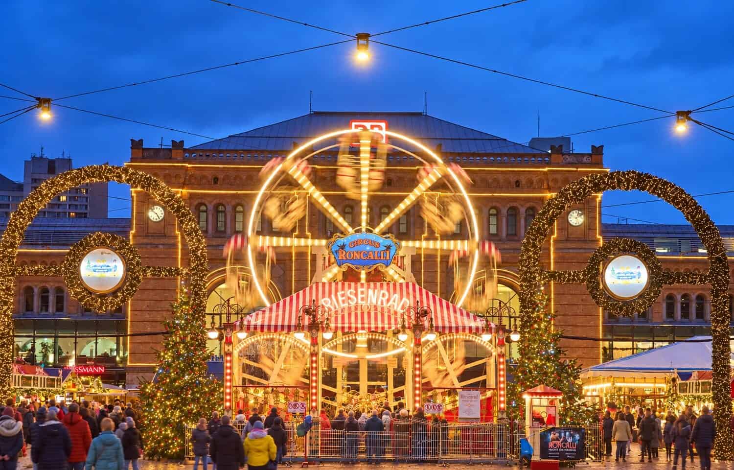 Beleuchtetes Riesenrad im Roncalli Weihnachtsdorf vor dem Hauptbahnhof Hannover zur blauen Stunde, davor Lichterbögen, Weihnachtsbäume und Besucher auf dem Ernst-August-Platz