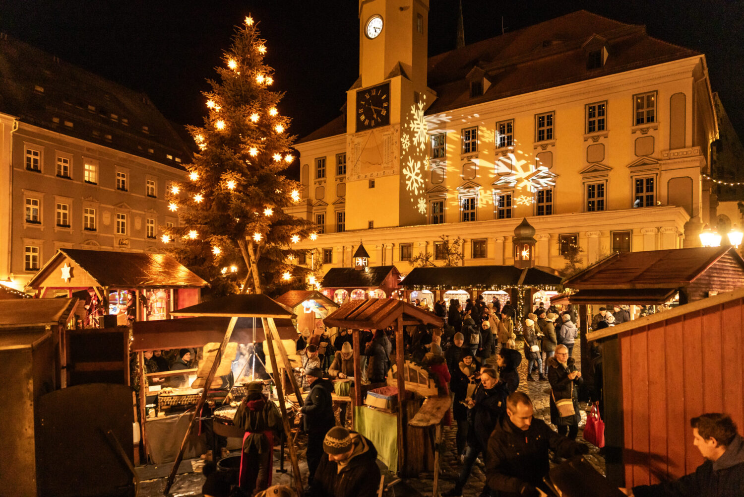 Besucher zwischen Holzbuden auf dem Bautzener Wenzelsmarkt, dahinter Weihnachtsbaum und Rathaus mit Schneeflocken-Projektion